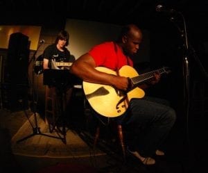 Joel Cross in red shirt on stage with his Benedetto Bravo jazz guitar