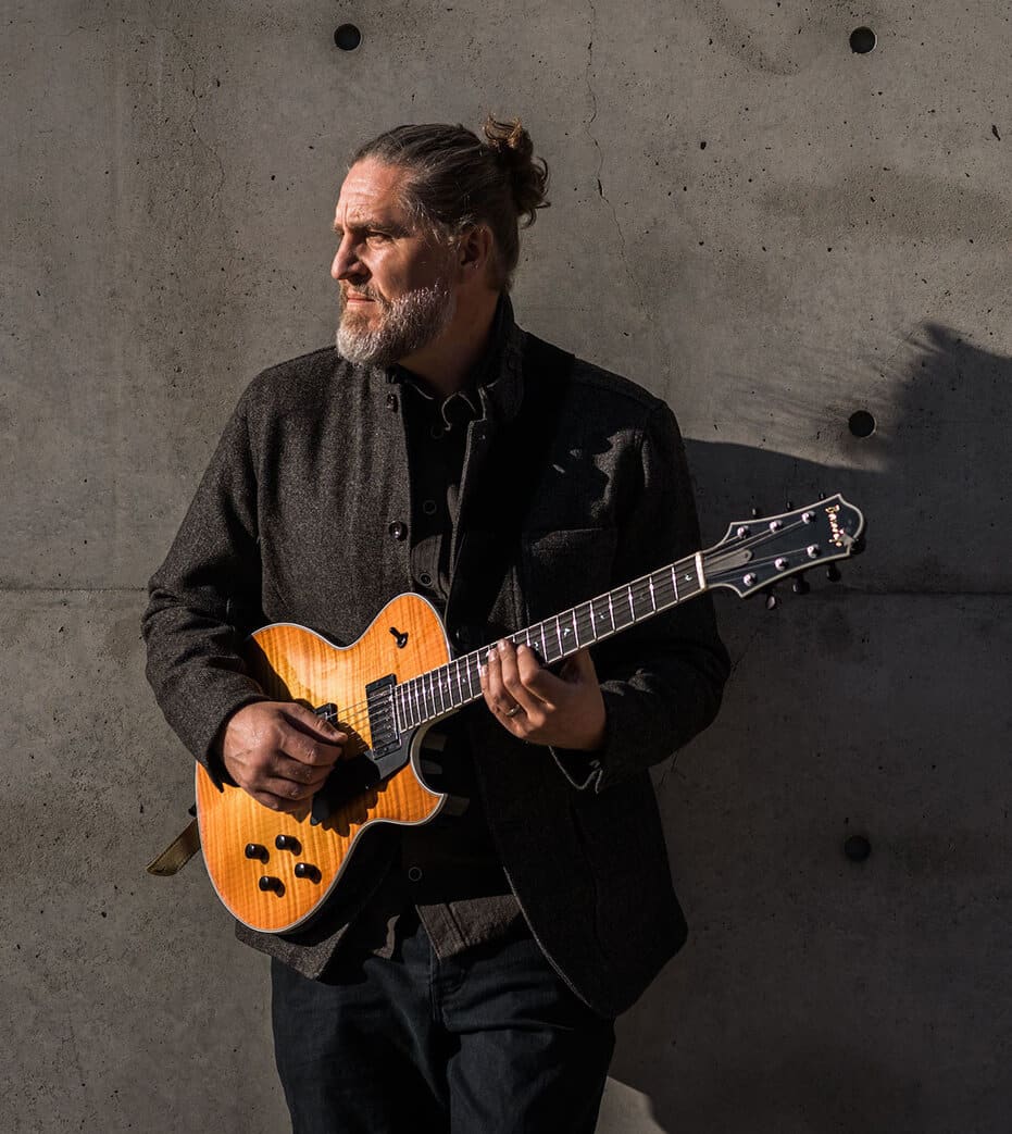 Corey Christiansen standing against a concrete wall playing a Benedetto Guitar.