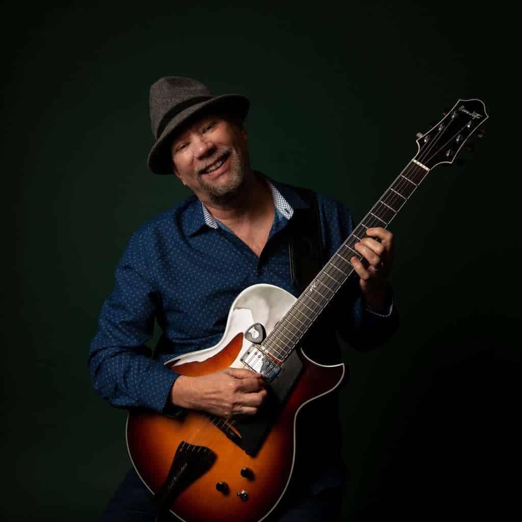 Paul Bollenback against a black background holding a Benedetto guitar.