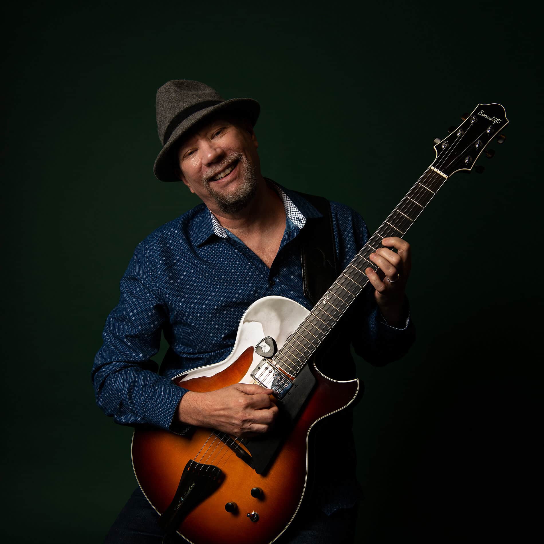 Paul Bollenback against a black background holding a Benedetto guitar.