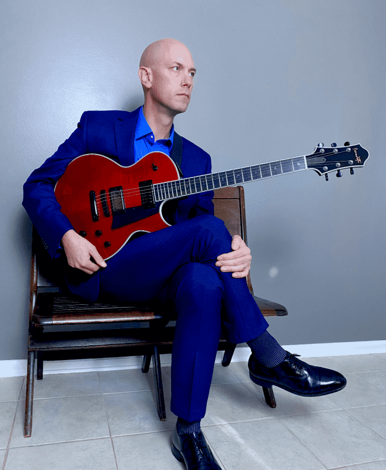 Shawn Purcell sitting down and holding a red Benedetto guitar.