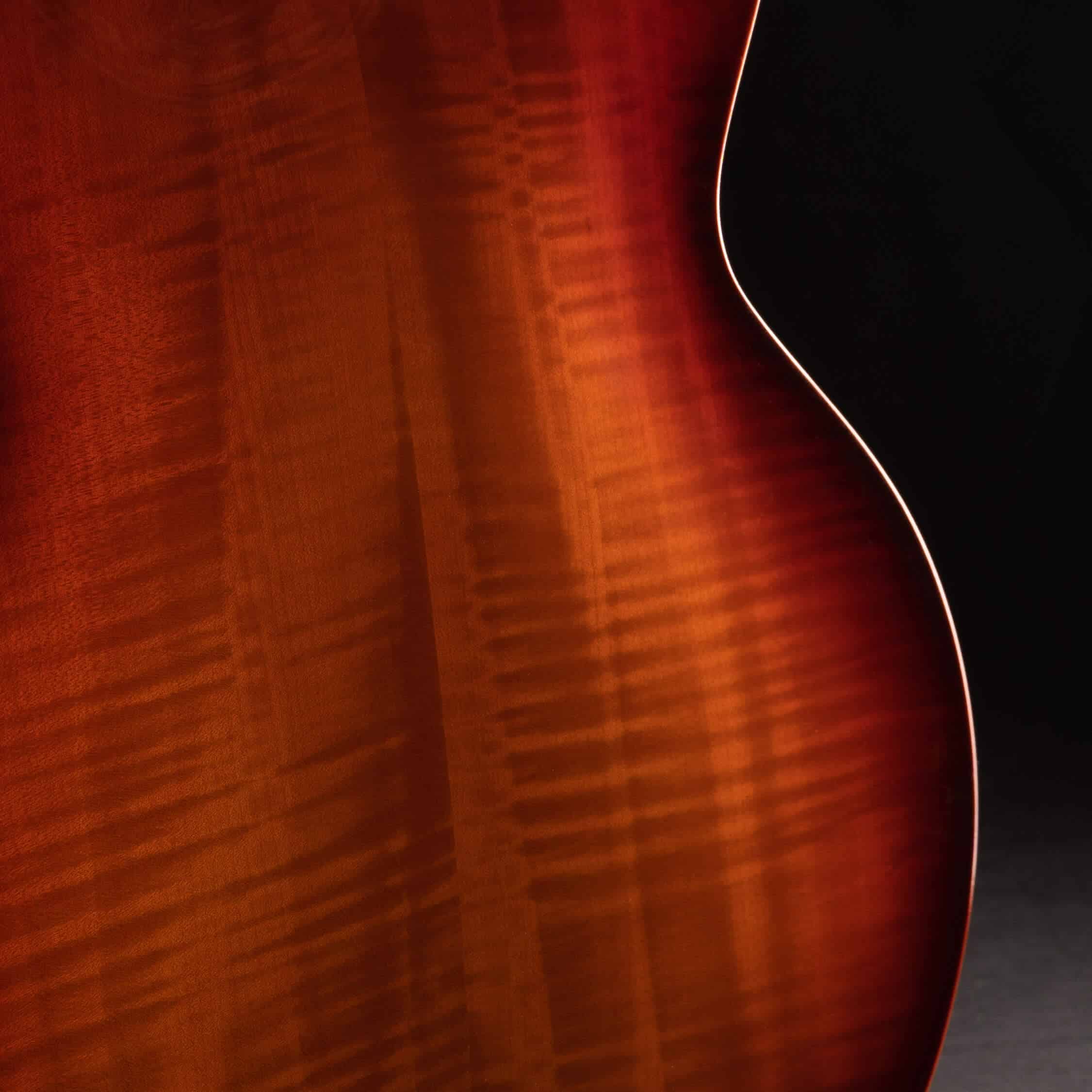 Macro photograph of a Benedetto Bravo Elite archtop guitar lower bout, showcasing highly figured flamed maple wood grain under a rich, high-gloss honey sunburst lacquer finish against a black background.