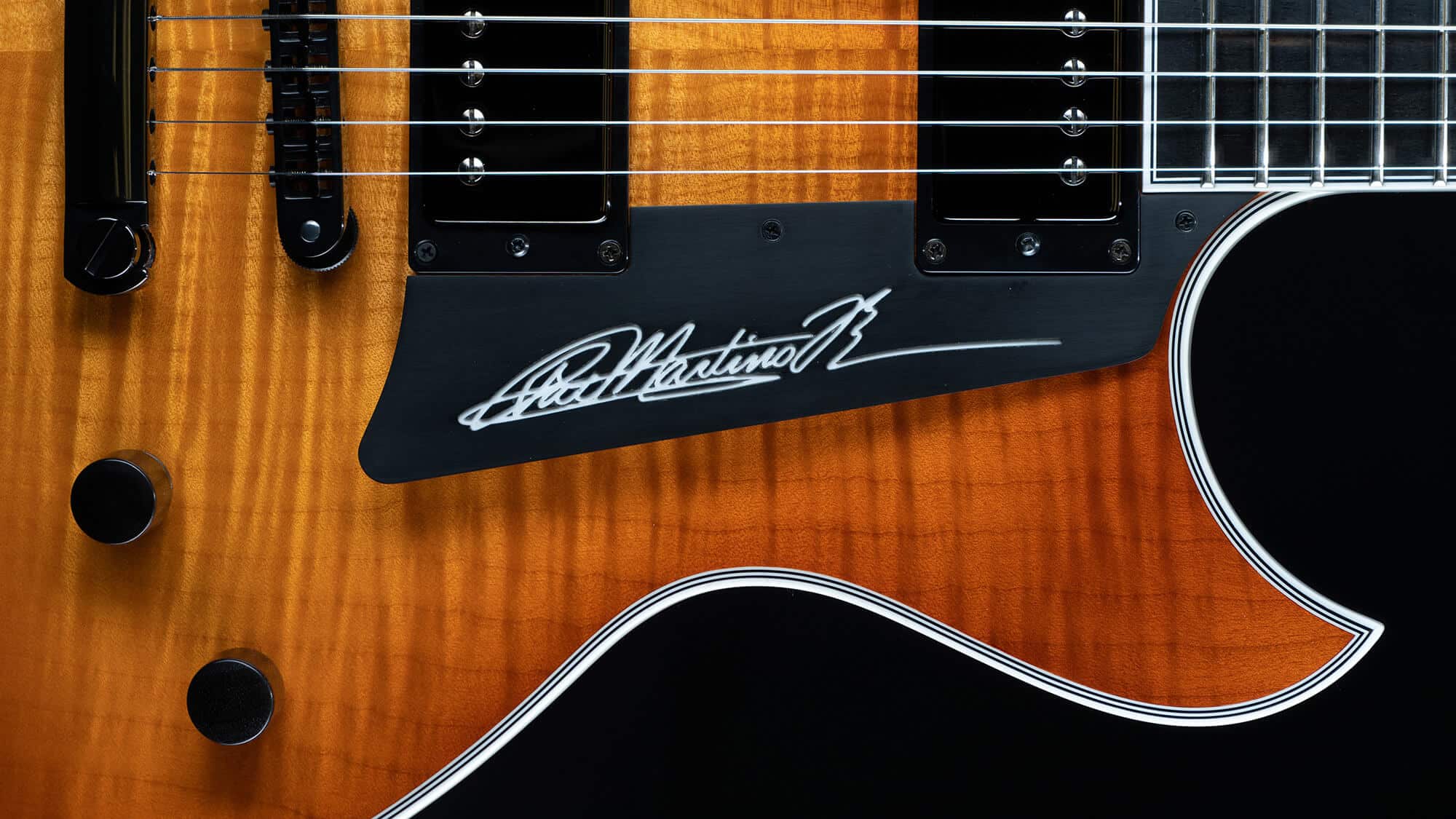 A detailed top-down view of the body of a maple-top Benedetto Pat Martino Signature Model guitar, showcasing the distinct dual humbucker pickups, control knobs, and Pat Martino's signature script elegantly inlaid onto the black pickguard.