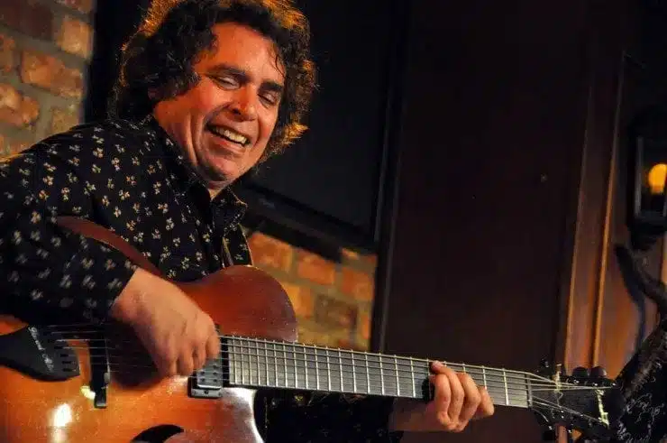 Jazz guitarist Howard Alden smiling widely while performing on stage, playing a sunburst-finish Benedetto archtop guitar against a background with a brick wall element.
