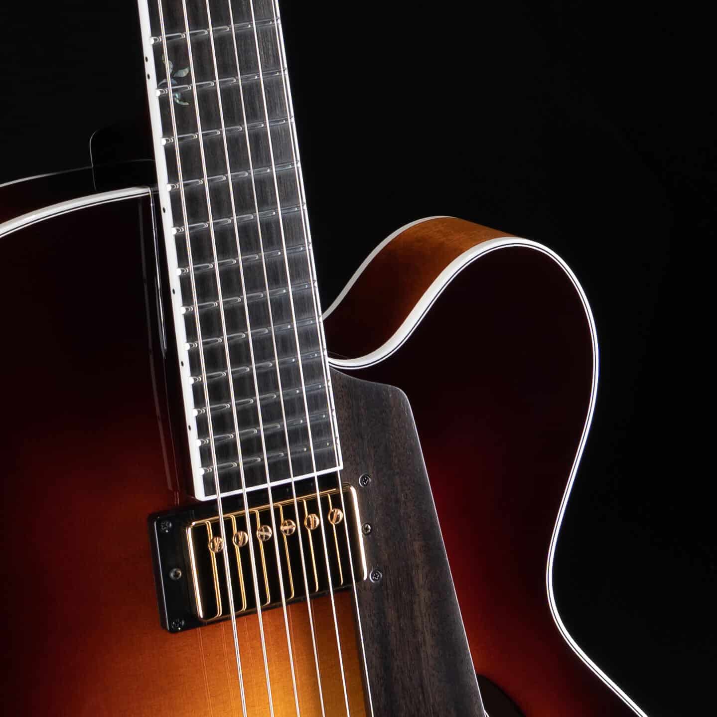 Close-up of the upper bout and neck joint of a sunburst Benedetto Bravo archtop guitar. Features an ebony fingerboard with a signature floral inlay at the 12th fret, a black humbucker pickup mounted adjacent to a matching grained ebony pickguard, and bright white binding contouring the Venetian cutaway.