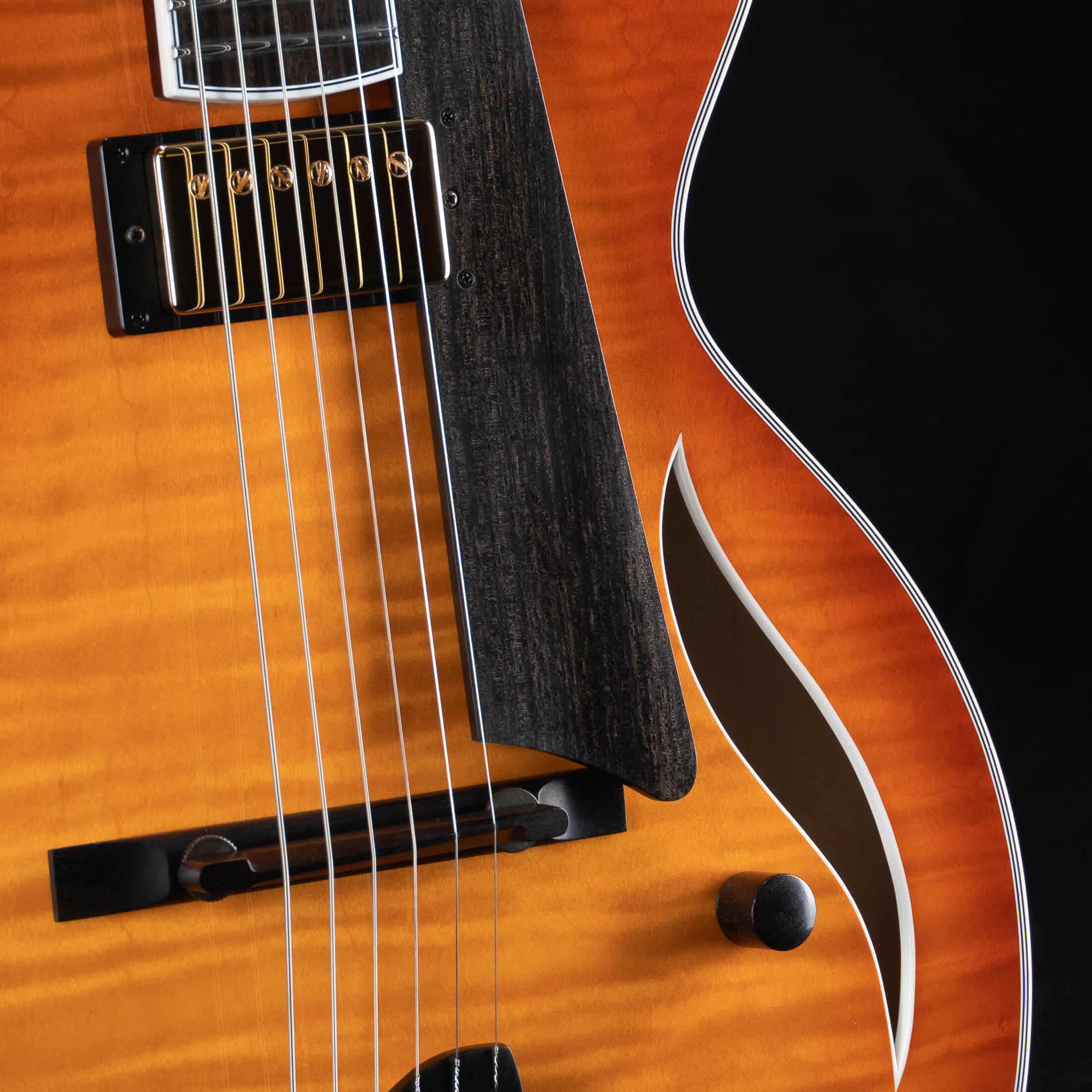 Close-up detail of the top soundboard of a Benedetto Bambino Deluxe archtop guitar in a vibrant honey sunburst finish over flamed maple. The shot highlights a black humbucker pickup with gold pole pieces, an organic-looking matte ebony pickguard and floating bridge, a stylized S-shaped soundhole, and a single ebony control knob.