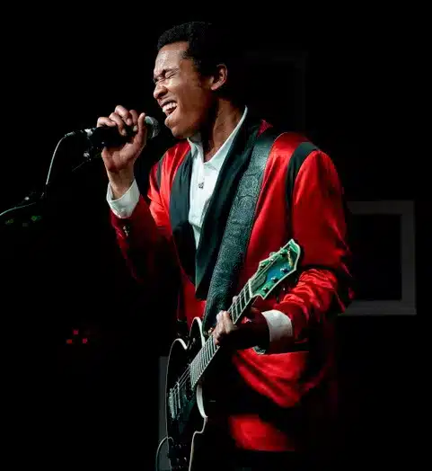Jazz and blues musician Solomon Hicks singing passionately into a microphone while holding his black Benedetto solid-body guitar on stage, wearing a striking red and black satin jacket over a white collared shirt.