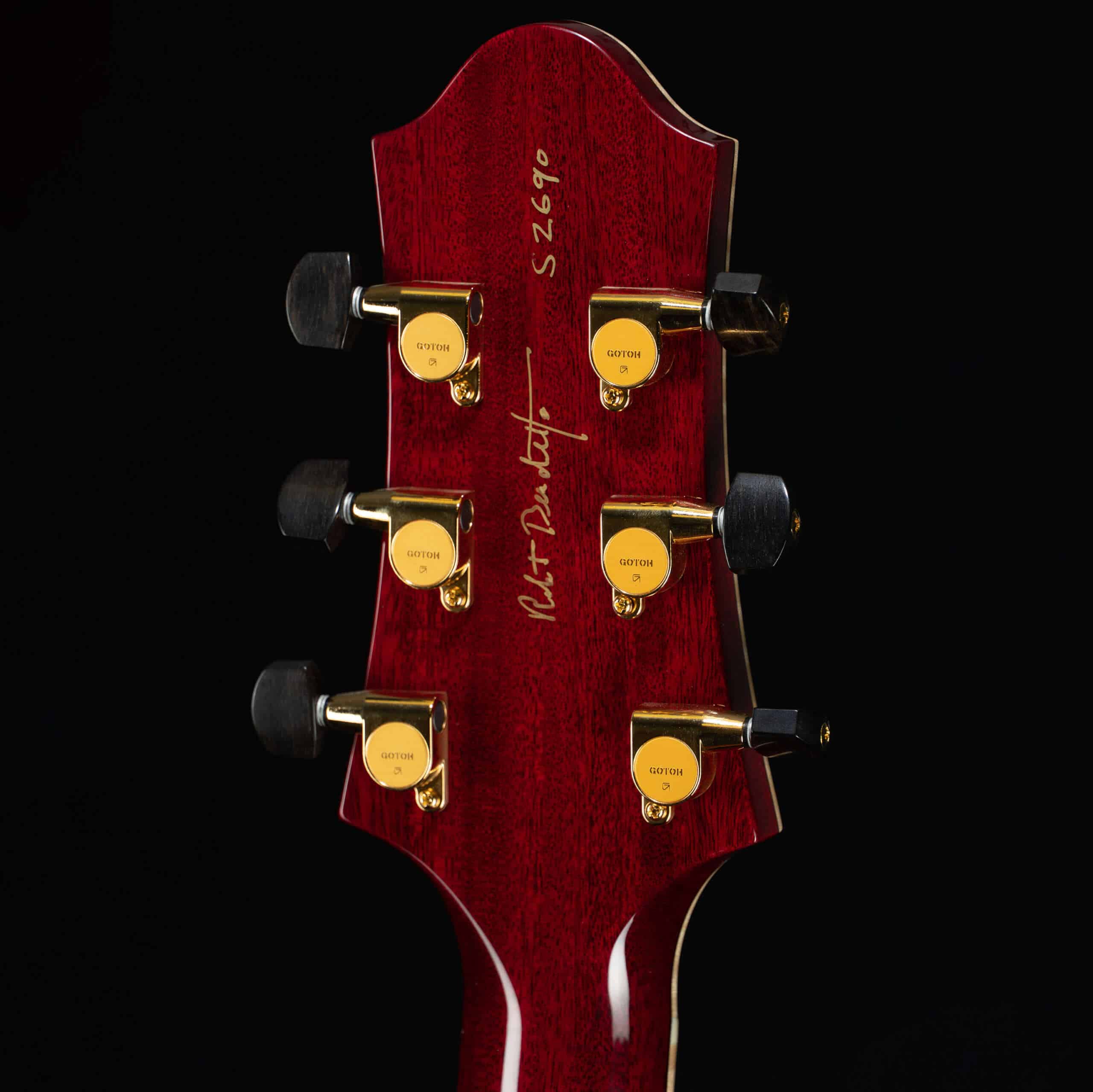 Close-up of the back of a Benedetto Benny model headstock with a rich, translucent ruby-red gloss finish over a subtle wood grain. The headstock features six gold Gotoh tuning machines with dark ebony buttons, a hand-written gold serial number "S2690" at the top, and Bob Benedetto's signature elegantly inscribed in gold ink down the center.