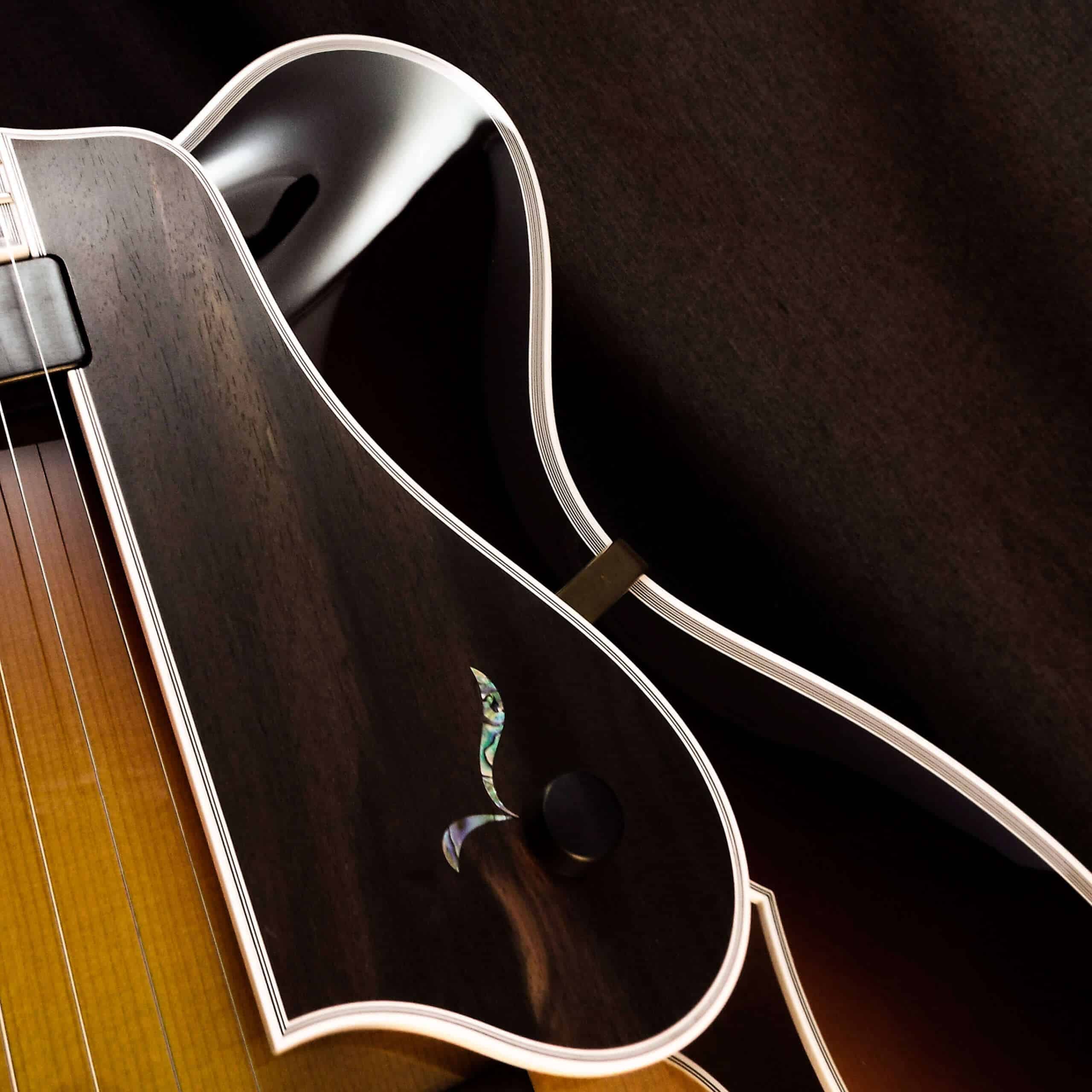Macro view of a Benedetto Cremona archtop guitar’s cutaway, showcasing an ebony pickguard inlaid with a colorful abalone shell floral accent. The shot highlights the meticulous white multi-layered binding tracing the pickguard, the tight grain of the spruce soundboard, and a glimpse of an iconic ebony f-hole edge in the background.