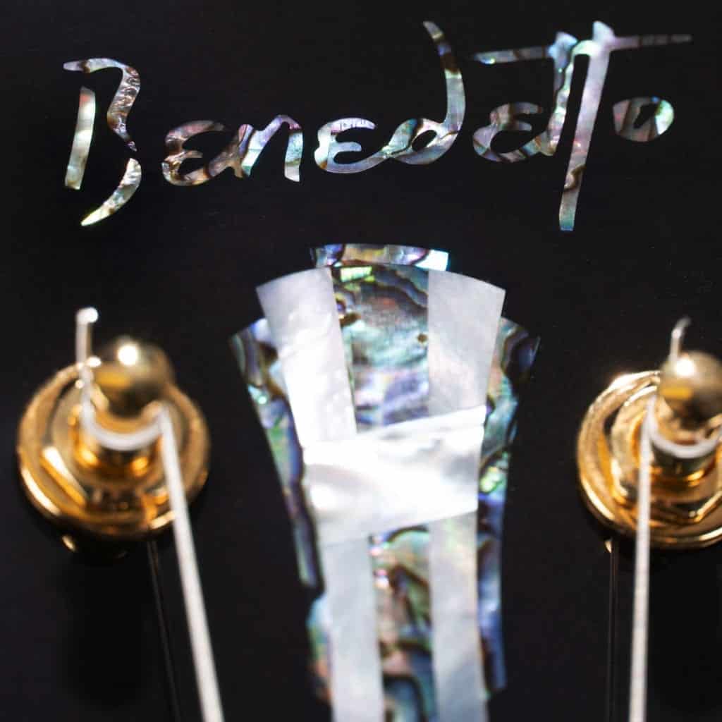 Close-up of a black guitar headstock featuring a pearl Benedetto script inlay and gold hardware.
